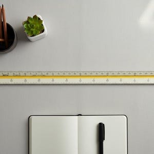 May include: A top-down view of a workspace with a ruler, pencils, a succulent, and a notebook. The ruler is yellow and white, with measurements in inches. The notebook is open, and a black pen rests on the right page. A black mug holds pencils.
