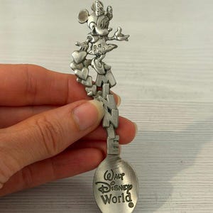 Może przedstawiać: Srebrzysta metalowa łyżeczka pamiątkowa z wyrzeźbioną figurką Myszki Minnie stojącą na pionowych literach tworzących napis MINNIE. Czerpak łyżeczki jest wygrawerowany czarnym napisem z logo Walt Disney World.