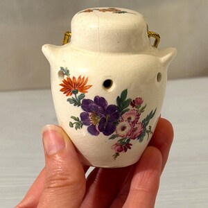 May include: A small, cream-coloured ceramic jar with a matching lid and a crackled glaze finish. The jar features a floral pattern with orange, purple, and pink flowers and green leaves. It has two small handles and a series of circular holes.