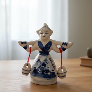 Könnte beinhalten: Keramikfigur einer holländischen Frau in traditioneller Tracht, bemalt in Weiß und Blau. Die Figur hält zwei kleine, gestreifte Behälter, die an roten Schnüren hängen. Das Kleid zeigt eine Windmühlenszene und das Wort "Holland".