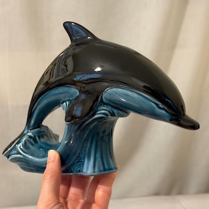 Könnte beinhalten: Eine glänzende Keramik-Delfinfigur mit einem glatten schwarzen Oberkörper und einem tiefblauen Bauch. Der Delfin ist in einer Sprungbewegung dargestellt und taucht aus einer stilisierten, texturierten blauen Wellenbasis auf.
