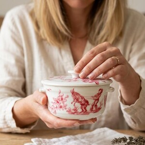 May include: A white ceramic casserole dish with a lid, featuring a red pastoral scene design. The dish has small handles and is held by a person. The design includes figures and animals. The dish is on a wooden surface.