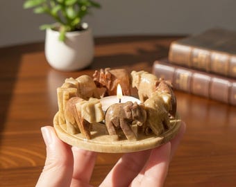 May include: A candle holder with a circular design, featuring carved elephants in shades of brown and beige. A lit candle sits in the center. The holder is held in a hand, with a plant and books in the background.