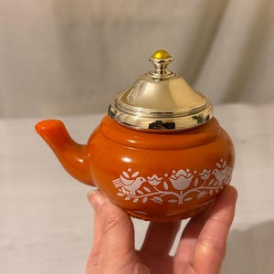 Könnte beinhalten: Ein Set aus drei Keramikbechern mit gesprenkelter, cremefarbener Glasur. Jeder Becher zeigt ein handgemaltes Design: ein blau-grünes Blatt, eine gelbe Blume und eine rote Beere. Die Becher haben eine einfache, abgerundete Form.