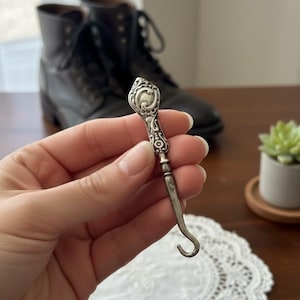 May include: A silver metal button hook with an ornate handle and a curved hook end. The hook is held in a hand, with a pair of black leather boots in the background. A small succulent plant is also visible.