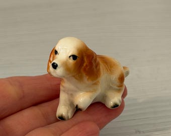 May include: A small ceramic figurine of a spaniel dog in a lying position. The dog has a cream-colored face and body with patches of light brown on its ears and back. It features painted black eyes, a black nose, and black tips on its paws.