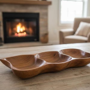 Könnte beinhalten: Eine Holzservierschale mit drei verbundenen Fächern. Die Schale hat eine natürliche Holzmaserung und ein glattes, geschwungenes Design. Die Schale steht auf einem hellen Holztisch.