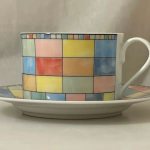 May include: A white teacup and saucer set with a colourful geometric design. The cup and saucer feature a grid pattern of pastel squares in shades of pink, yellow, blue, and green. The cup has a curved handle.