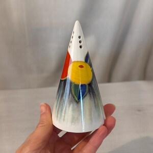 May include: A white, cone-shaped salt shaker with a colorful floral design. The design includes red, yellow, blue, and green accents. The shaker has three holes near the top and is held in a hand.
