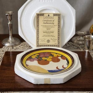 May include: A decorative plate with a floral design in a protective foam case. The plate has a yellow border and a colorful floral pattern. A certificate of authenticity is included. The plate is on a wooden surface.