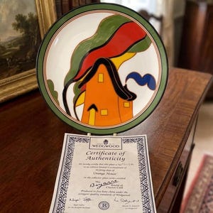 May include: A decorative plate with a colorful design featuring an orange house, green foliage, and red and yellow accents. The plate is displayed with a certificate of authenticity from Wedgwood.