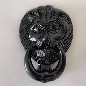 Könnte beinhalten: Ein schwarzer Türklopfer in Löwenform mit ringförmigem Griff. Der Klopfer weist detaillierte Merkmale auf, darunter Mähne, Augen, Nase und Bart. Der Türklopfer besteht aus glänzendem Material und ist auf einer weißen Oberfläche montiert.