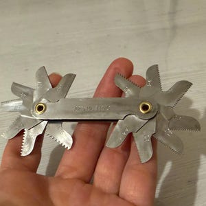 Könnte beinhalten: Eine silberfarbene Gewindelehre aus Metall mit mehreren klappbaren Blättern mit gezackten Kanten. Der mittlere Griff ist mit dem Text 'AF-METRIC' geprägt und verfügt über zwei messingfarbene Nieten, die die Blätter an beiden Enden sichern.