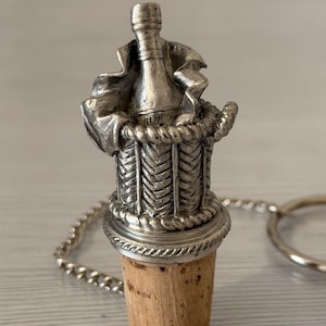 May include: Silver-toned bottle stopper featuring a champagne bottle design within a basket. The stopper has a cork base and a decorative chain with a ring. A unique wine accessory.