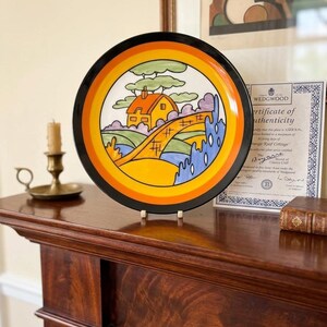 May include: A decorative plate with a black rim and a colorful illustration of a house and landscape. The plate has a yellow, orange, and blue color scheme. The plate is displayed on a wooden surface.