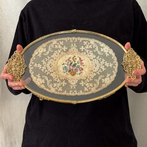 以下が含まれることがあります： 金色の金属フレームと両側に装飾的なフィリグリースタイルのハンドルを備えた楕円形のサービングトレイ。トレイの中央には、赤、青、緑の色調で刺繍された花のブーケがあしらわれたクリーム色のレースが配置されています。