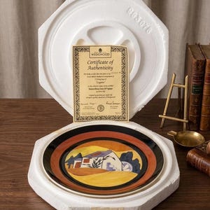 May include: A decorative plate with a colorful landscape design, featuring a house and trees, is nestled in a protective foam insert. The plate has a black and orange border. A certificate of authenticity is visible.