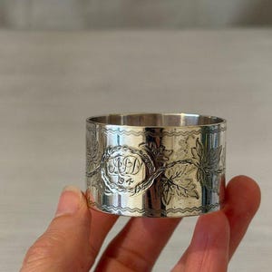 May include: A cylindrical silver napkin ring featuring an engraved pattern of leaves and vines. The centre of the design displays an oval frame containing the engraved letters "AJD" positioned above the number "94". The edges have a zigzag border.