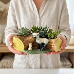 Könnte beinhalten: Ein Keramik-Pflanzgefäß in Form eines Baumstamms mit einem Lamm und einem Hasen, gefüllt mit Sukkulenten. Der Pflanztopf hat ein hellbraunes und gelbes Farbschema mit grünen Akzenten. Das Lamm hat schwarze Beine und einen weißen Körper.