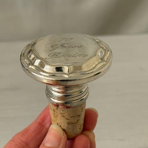 Pode incluir: Uma rolha de garrafa prateada com base de cortiça. A parte superior da rolha está gravada com as palavras "70 Years Ronine". A rolha tem uma borda decorativa e canelada.