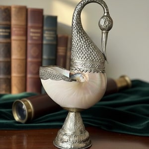 May include: A decorative nautilus shell with ornate silver-colored metalwork. The shell is mounted on a pedestal base and features a curved, swan-like neck. The background includes antique books and a telescope.