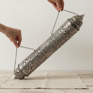 Puede incluir: Un recipiente cilíndrico plateado con intrincados patrones grabados y una parte superior puntiaguda. El recipiente está suspendido por cadenas y sostenido por manos, lo que sugiere que es un artículo decorativo o ceremonial. El artículo descansa sobre una tela de lino beige.
