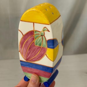 May include: A decorative ceramic salt shaker with a yellow top and a rectangular body. The body is painted with a colorful design of a fruit and a bow. The base has blue feet. The shaker is held in a hand.