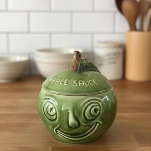 May include: Green ceramic apple-shaped jar with a smiling face design. The lid is inscribed with "APPLE SAUCE". The jar has a brown stem and green leaf detail. The jar is sitting on a wooden surface.