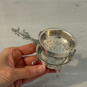Könnte beinhalten: Ein Teesieb aus silberfarbenem Metall mit einer perforierten Schale und einem dekorativen Griff, der eine skulptierte Figur zeigt. Das Sieb ruht auf einem passenden runden Sockel mit drei kleinen Füßen, die jeweils oben ein muschelartiges Design aufweisen.