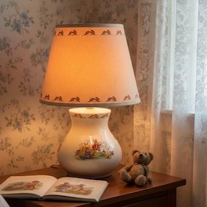 Può includere: Una lampada da tavolo decorativa con un paralume beige con un motivo a conigli ripetuto. La base in ceramica presenta una scena pastorale. Un piccolo orsacchiotto e un libro aperto sono sul comodino di legno.