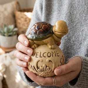 May include: A ceramic turtle ornament with a brown and tan shell, sitting on a round base. The base is tan with the word "WELCOME" and floral designs. The ornament is held in someone's hands.