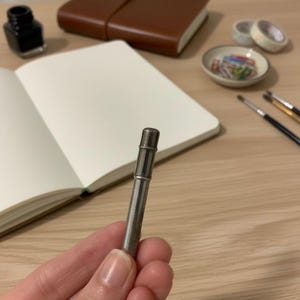 Puede incluir: Un portaminas plateado se sostiene frente a un cuaderno abierto. Otros artículos de papelería incluyen un diario de cuero marrón, un tintero, cinta washi, clips y pinceles. Los artículos están sobre una superficie de madera clara.