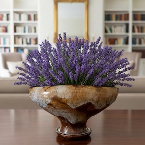 May include: A ceramic bowl filled with purple lavender, set on a dark wooden table. The bowl has a distinctive, earthy design with brown and cream tones. The background features a gold mirror and bookshelves, creating a warm atmosphere.
