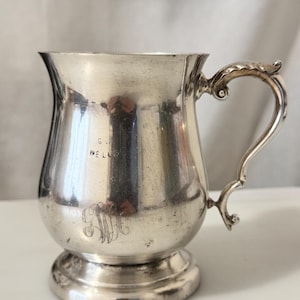May include: A silver-coloured mug with a curved handle and a decorative flourish at the top. The mug has a rounded body and a small base. The initials "GM" are engraved on the front, with the name "WELLS" above. The mug is likely vintage.
