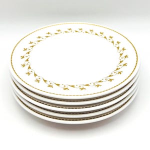 May include: A stack of five white ceramic plates. Each plate features a gold floral design around the rim. The plates have a scalloped edge and a simple, elegant aesthetic.