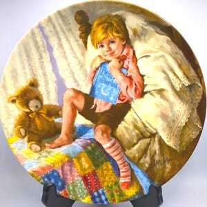 May include: Decorative plate featuring a young child holding a book titled "Bear Stories." The child is seated on a patchwork quilt with a teddy bear nearby. The plate has a soft, warm color palette.