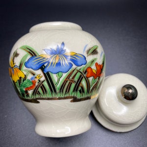 May include: A cream-colored ceramic jar with a lid, decorated with colorful floral and bird motifs. The jar features a blue iris, orange and yellow flowers, and a small bird. The lid has a silver knob.
