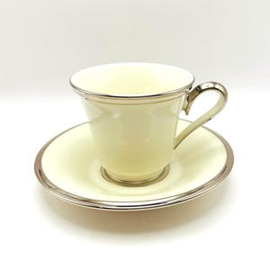 May include: A cream-colored teacup and saucer set with silver-colored trim. The teacup has a curved handle and sits on a matching saucer. The set is likely made of porcelain or a similar material, and is suitable for serving tea or coffee.