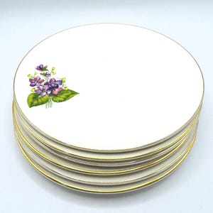 May include: A stack of white ceramic plates with a gold rim. Each plate features a floral design of purple violets and green leaves. The plates are stacked on top of each other.