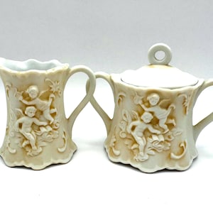 May include: A cream-colored ceramic creamer and sugar bowl set. The set features a detailed relief design of cherubs and floral accents. The creamer has a curved handle and a ruffled rim. The sugar bowl has a lid with a small circular handle.