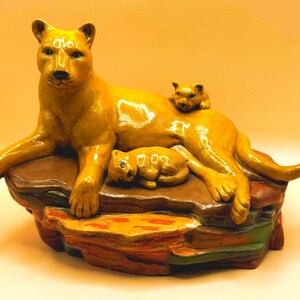 May include: A ceramic figurine of a golden-yellow lioness lying on a multi-colored rock formation. One small cub rests against the lioness's side, while another cub is positioned on her back. The base features brown, orange, and green tones.