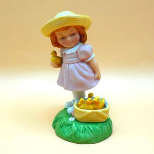 May include: A ceramic figurine of a young girl in a pink dress and yellow hat, holding a yellow duckling. A basket of yellow ducklings sits on a patch of green grass. The figurine has a vintage aesthetic.