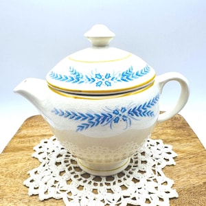 May include: Cream-colored ceramic teapot with a lid, featuring blue floral patterns and a yellow trim. The teapot has a curved spout and a handle. It sits on a wooden surface with a white lace doily.