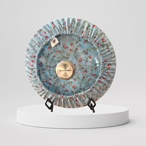 May include: A blue and white floral patterned fabric cover for a Le Creuset pot lid. The cover has a drawstring closure and a small tag with the Le Creuset logo.