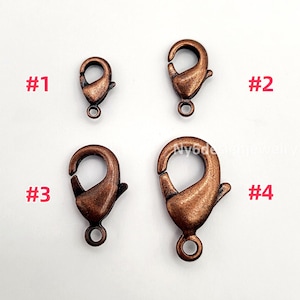 May include: Four antique copper-colored lobster claw clasps of varying sizes are displayed. Each clasp has a spring-loaded mechanism and a small loop for attaching to jewelry. The clasps are labeled with numbers 1 through 4.