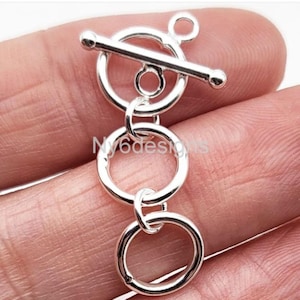 May include: A silver-colored toggle clasp with a circular design and a bar closure. The clasp is composed of multiple interconnected rings and a small loop. The image includes the text "Ny6designs".