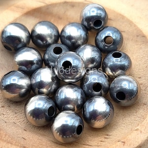 May include: A collection of shiny, silver-colored metal beads with a hole drilled through each. The beads are spherical and have a smooth, reflective surface. The beads are scattered on a wooden surface.