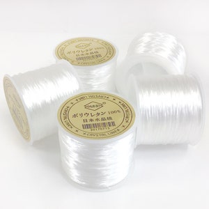 May include: Three spools of white elastic thread with a gold label that reads "ポリウレタン 100% 日本水晶线 20170713 CRYSTAL LINE".