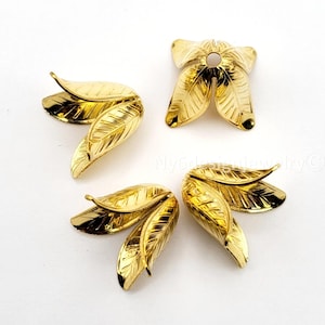 May include: Four gold-colored leaf-shaped jewelry findings on a white background. Each finding has detailed veining and a glossy finish. The findings are arranged in a circular pattern, with one centered and the others around it.
