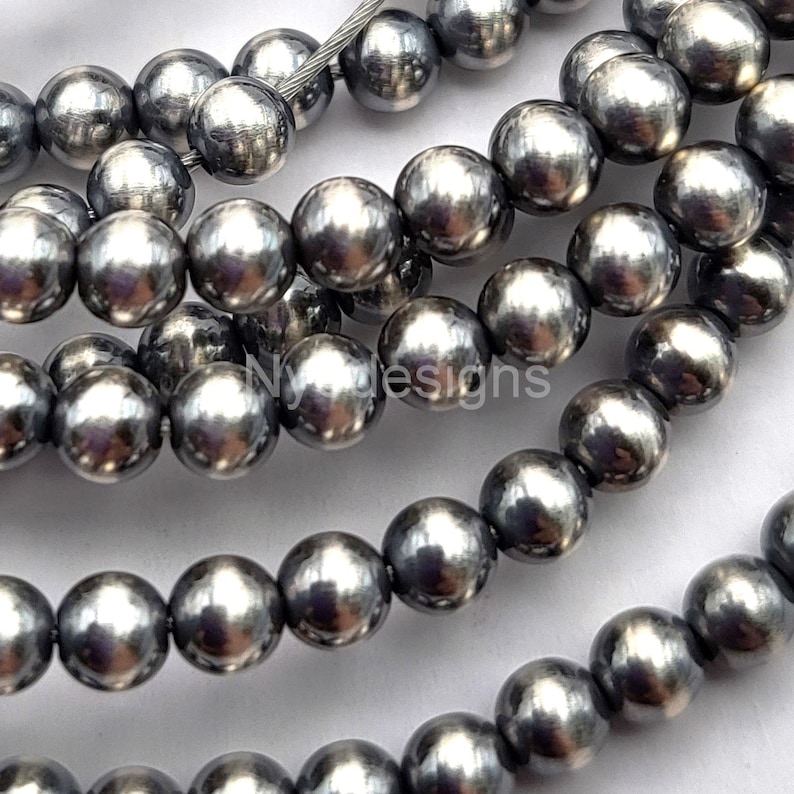 May include: Strands of metallic gray beads. The beads are spherical and have a shiny, reflective surface. The text "Ny designs" is visible on the beads. These beads are suitable for jewelry making.